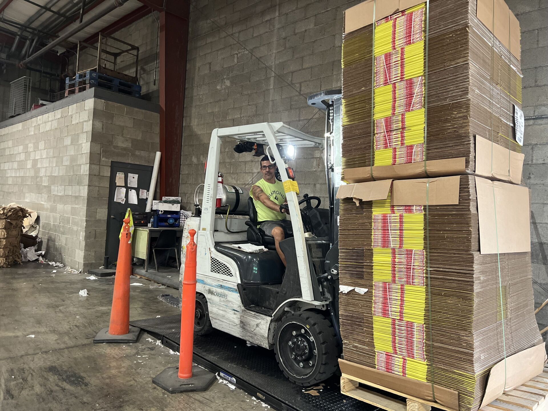 A man is driving a forklift in a warehouse next to a stack of cardboard boxes.