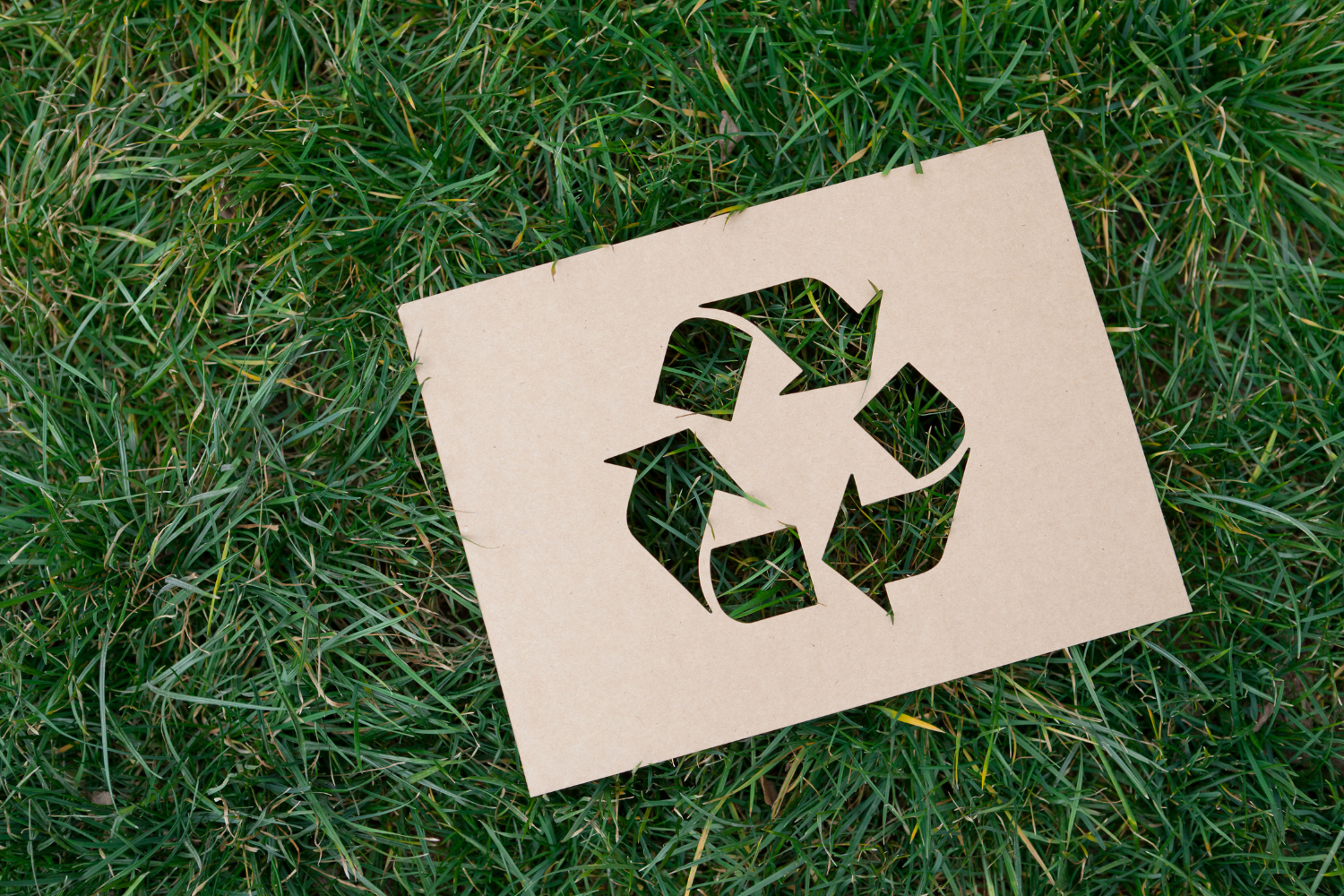 A cardboard recycling symbol is sitting on top of a lush green field of grass.