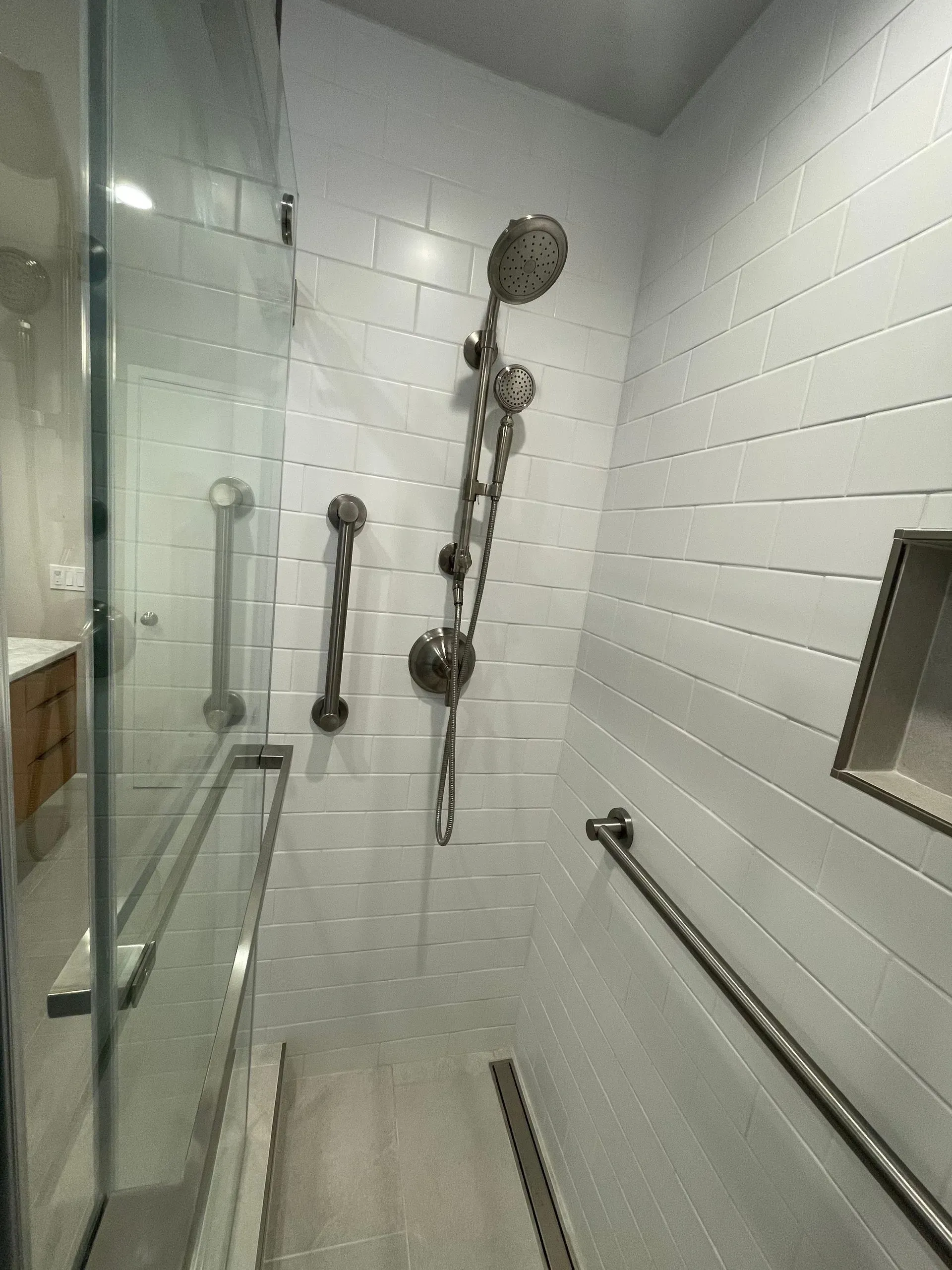 Glass shower with white tile walls and multiple chrome grab bars and shower fixtures