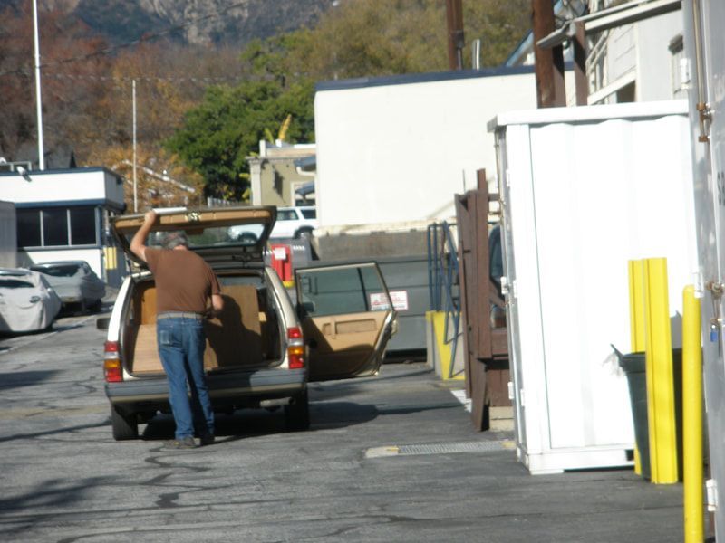 Man unloading boxes from a station wagon in a parking area with buildings and a mountain in the background.