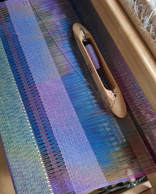 Weven met gekleurde draden op een weefgetouw. Het garen op de schietspoel is paars, op gedeeltelijk geweven stof zijn blauw- en paarstinten te zien.