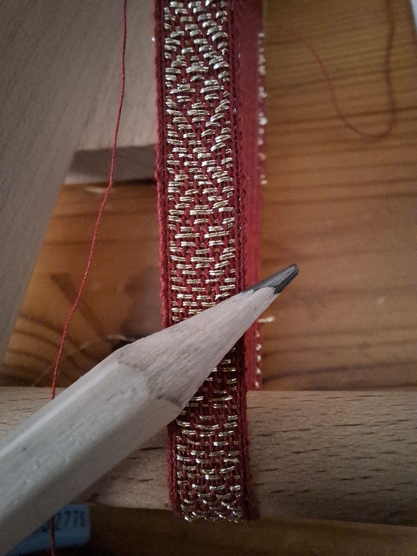 Potlood wijst naar de rode en zilveren geweven rand. De rand is vastgespeld op een houten frame; de rode draad is zichtbaar.