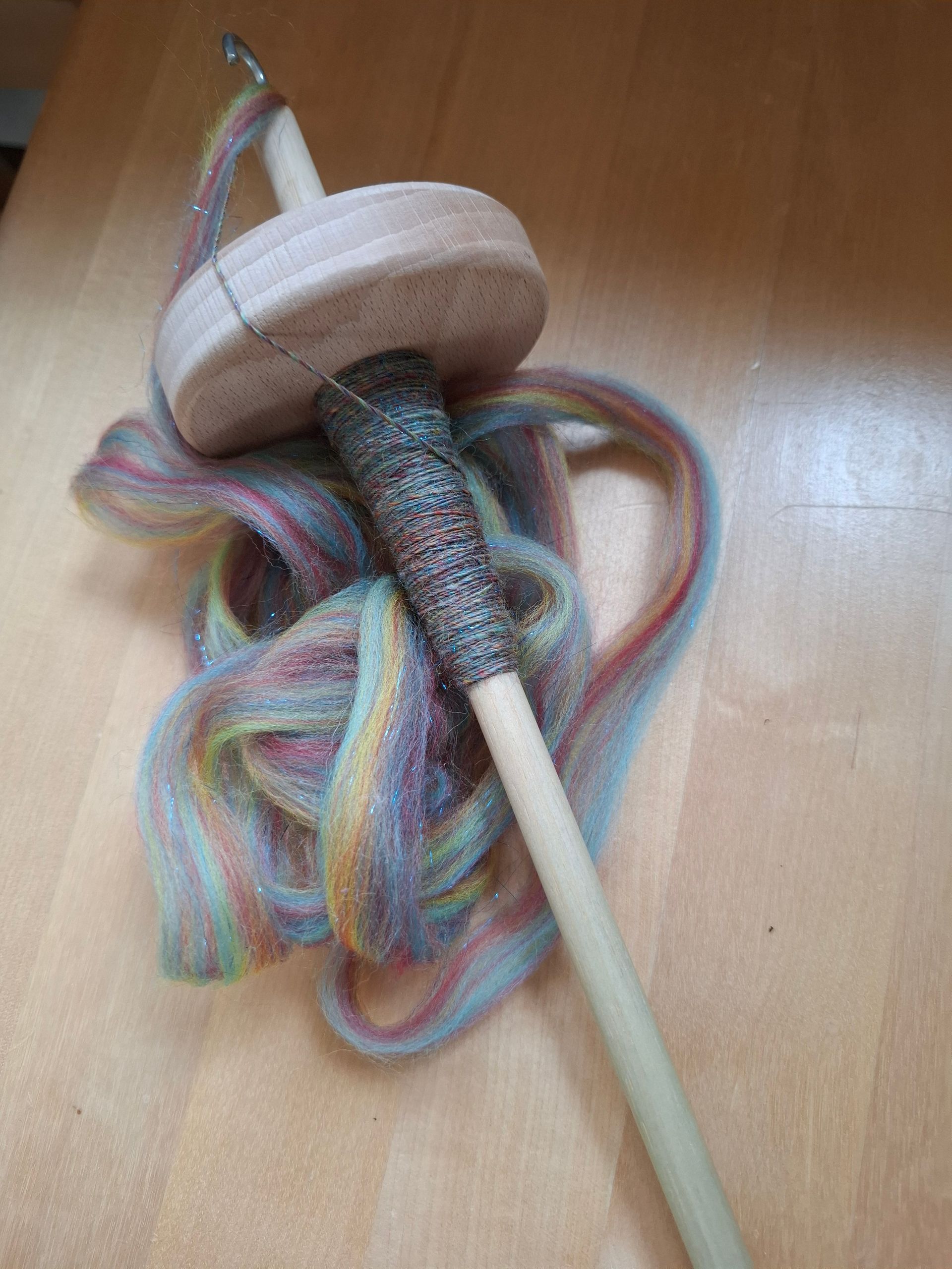 Een handspindel met gekleurde vezels eromheen gewikkeld, op een houten oppervlak.