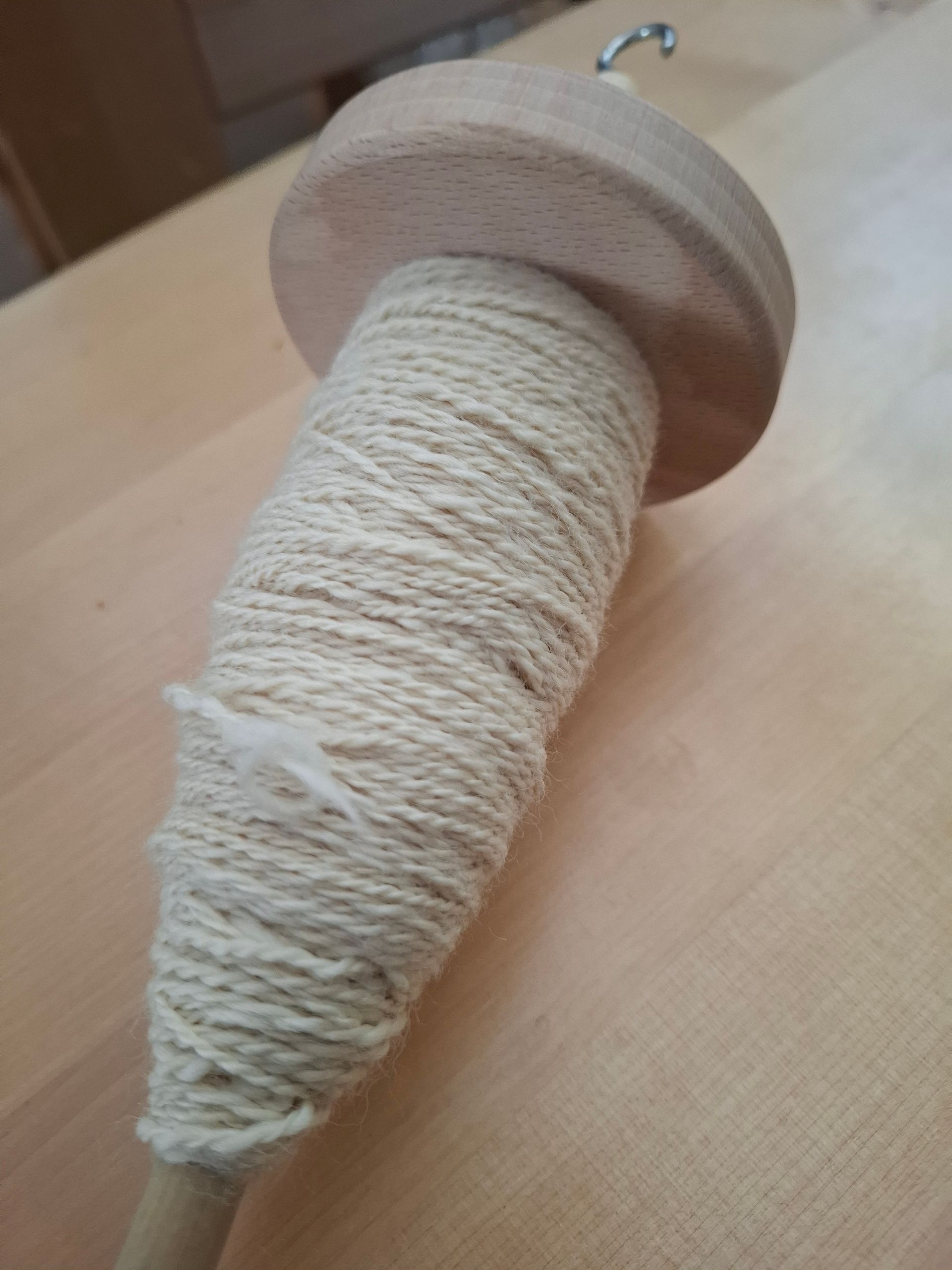 Een houten spintol, omwikkeld met crèmekleurig garen, ligt op een lichtgekleurd houten oppervlak.