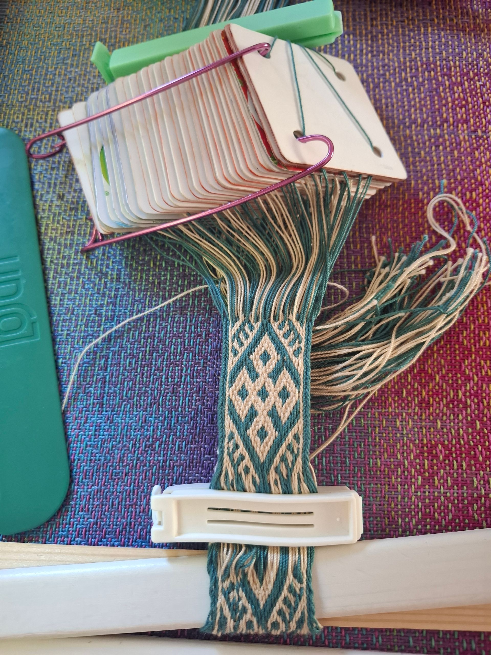 Weefproces van kaarten in uitvoering: blauwgroene en crèmekleurige band met geometrisch patroon. Kaarten, pinnen en gereedschap zichtbaar. Backstrap weven.