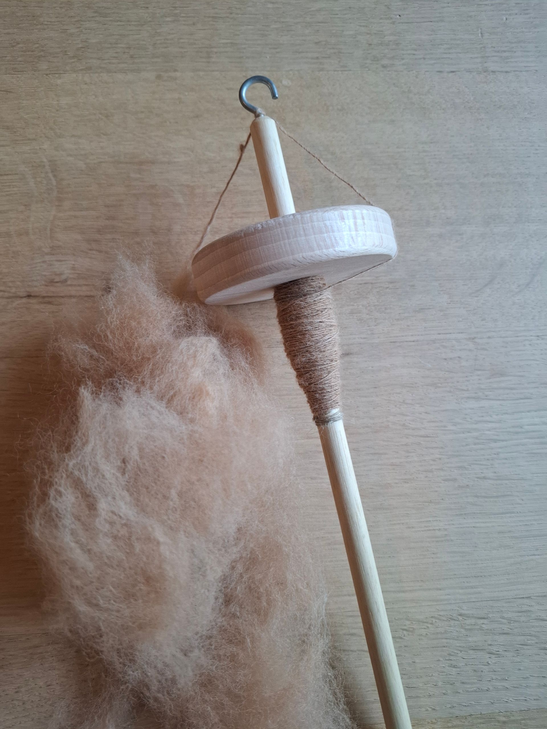 Een spintol met bruine wol. De spintol is van hout met een krans en haak. De wol ligt op het houtoppervlak.