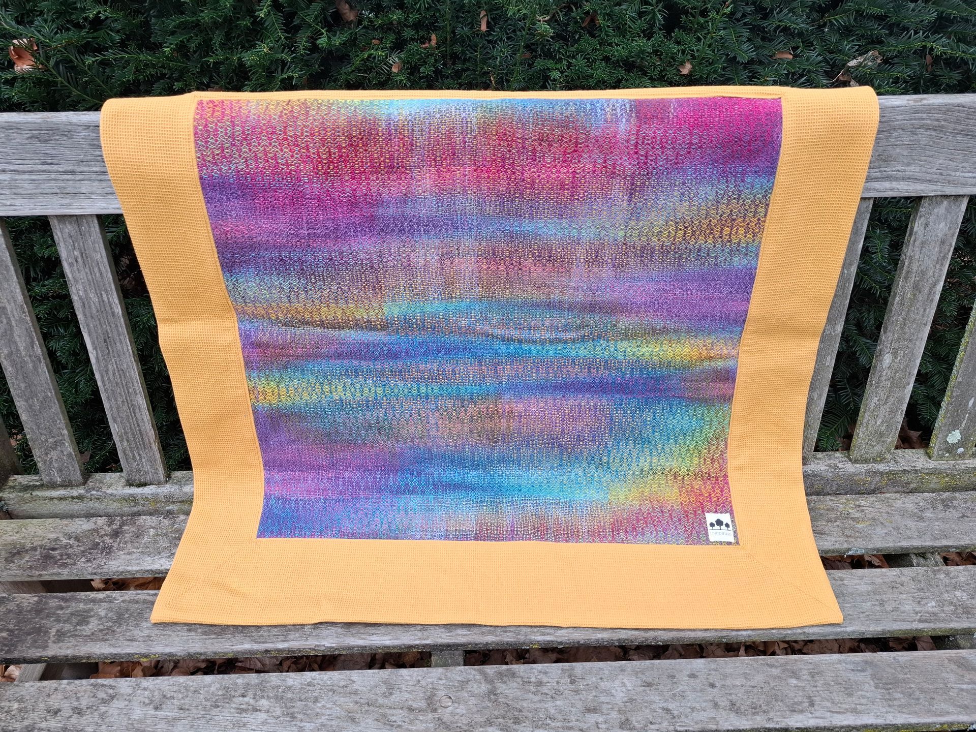 Kleurrijk textielkunstwerk met een mosterdgele rand op een houten bank.