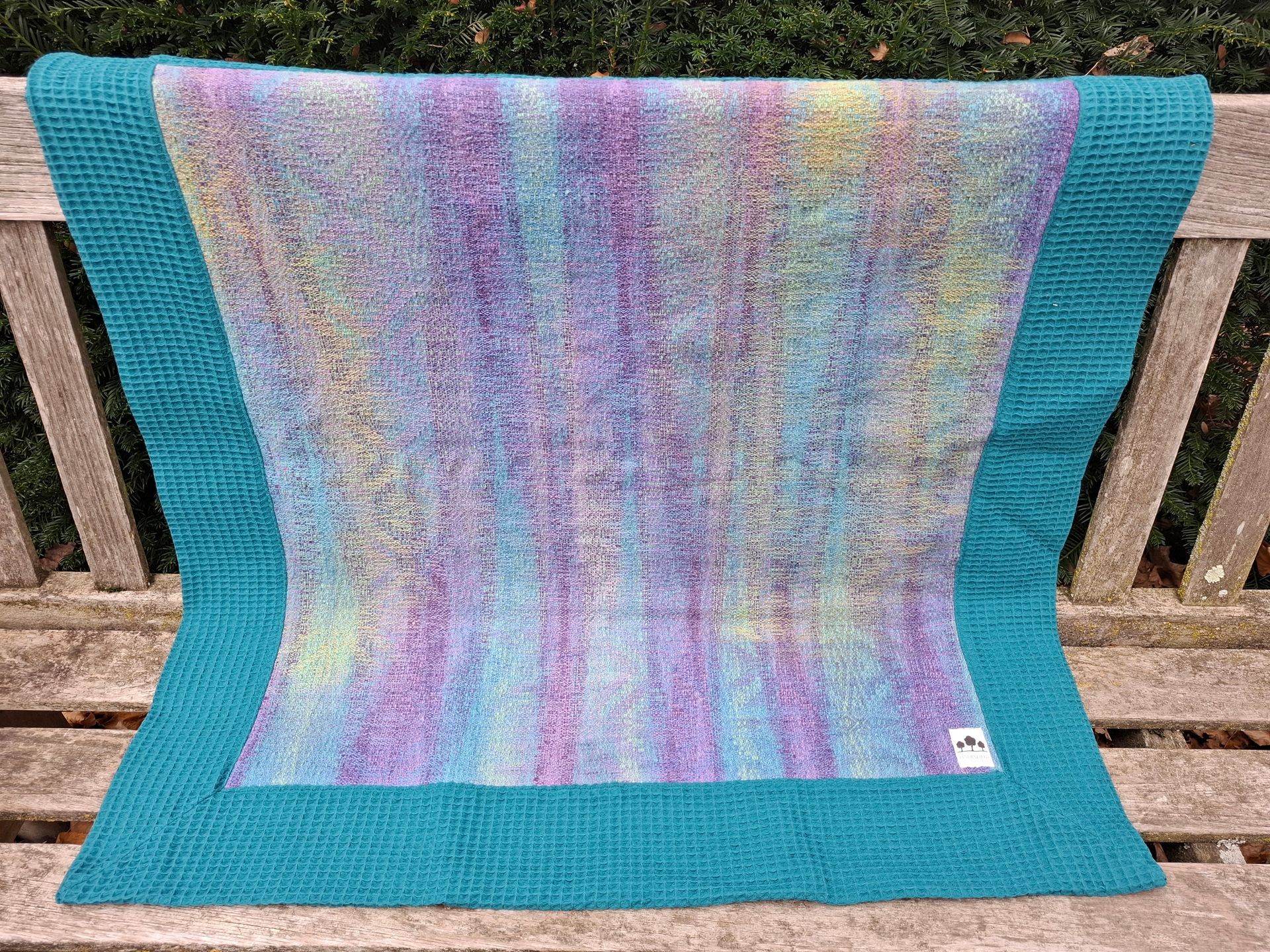 Een deken met een blauwgroene rand en een kleurrijk, verticaal strepenpatroon, gedrapeerd over een houten bank.