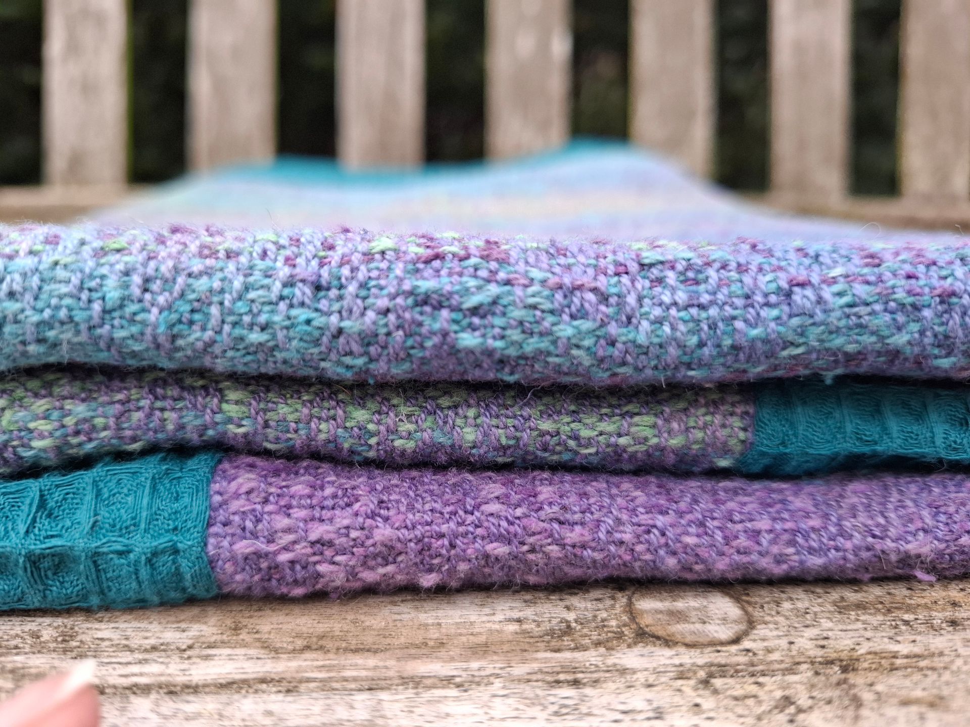 Een deken met een blauwgroene rand en een kleurrijk, verticaal strepenpatroon, gedrapeerd over een houten bank.