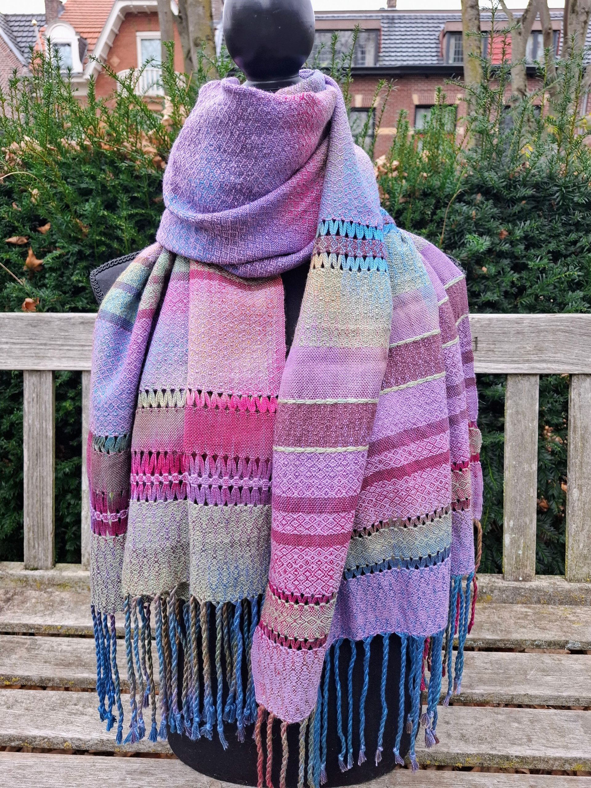 Een kleurrijke geweven sjaal hangt over een zwarte paspop, rustend op een houten bankje buiten.