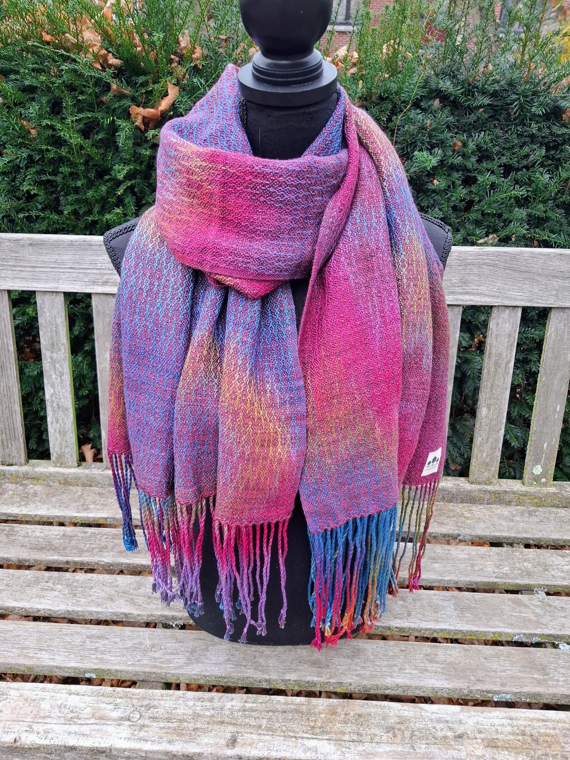 Kleurrijke geweven sjaal met franjes aan de uiteinden, gedrapeerd over een zwarte paspop op een houten bank.