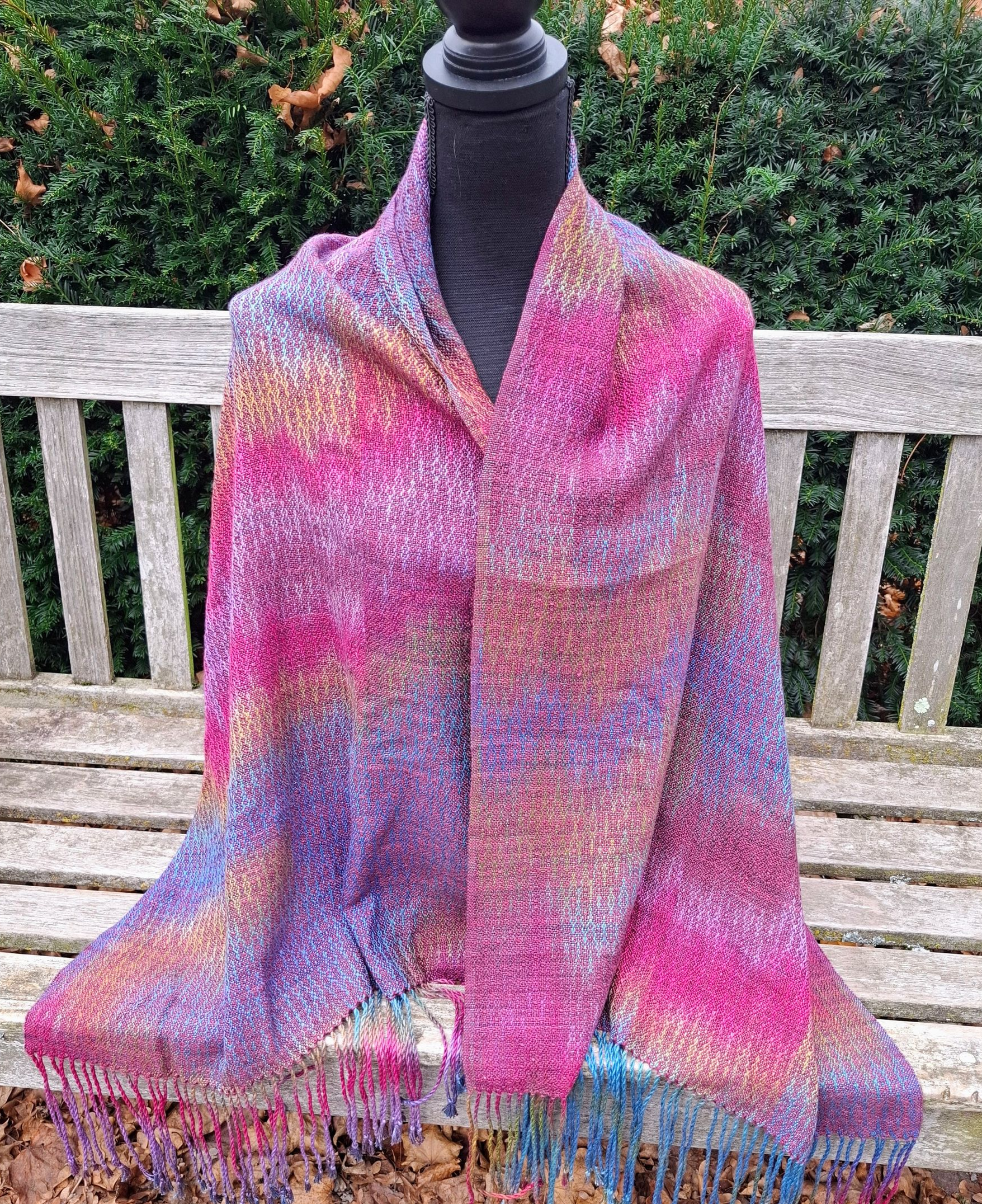 Kleurrijke geweven sjaal met franjes aan de uiteinden, gedrapeerd over een zwarte paspop op een houten bank.