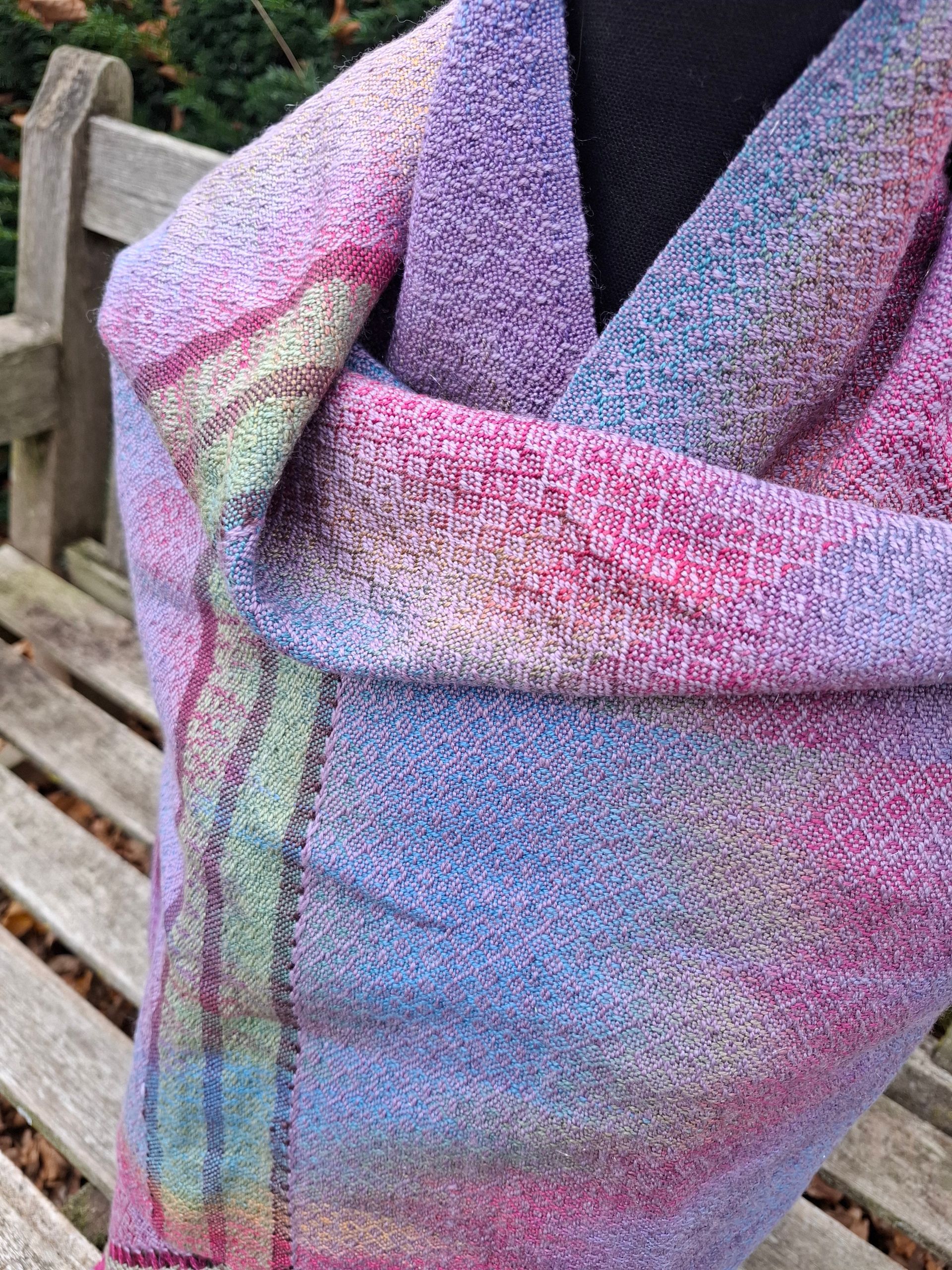 Een kleurrijke geweven sjaal hangt over een zwarte paspop, rustend op een houten bankje buiten.