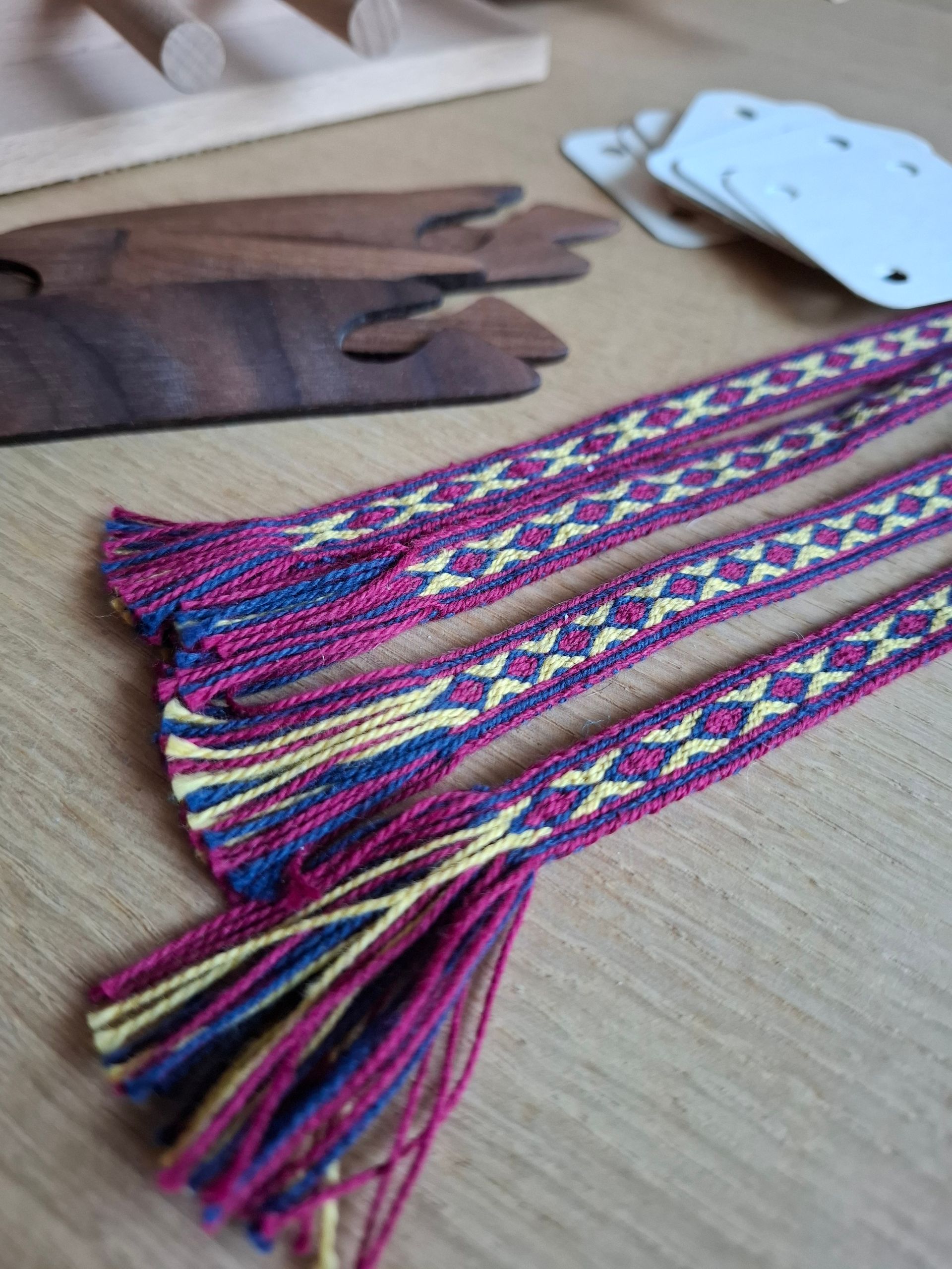 Drie geweven banden met een paars, blauw en geel patroon, met weefgereedschap op een houten oppervlak.