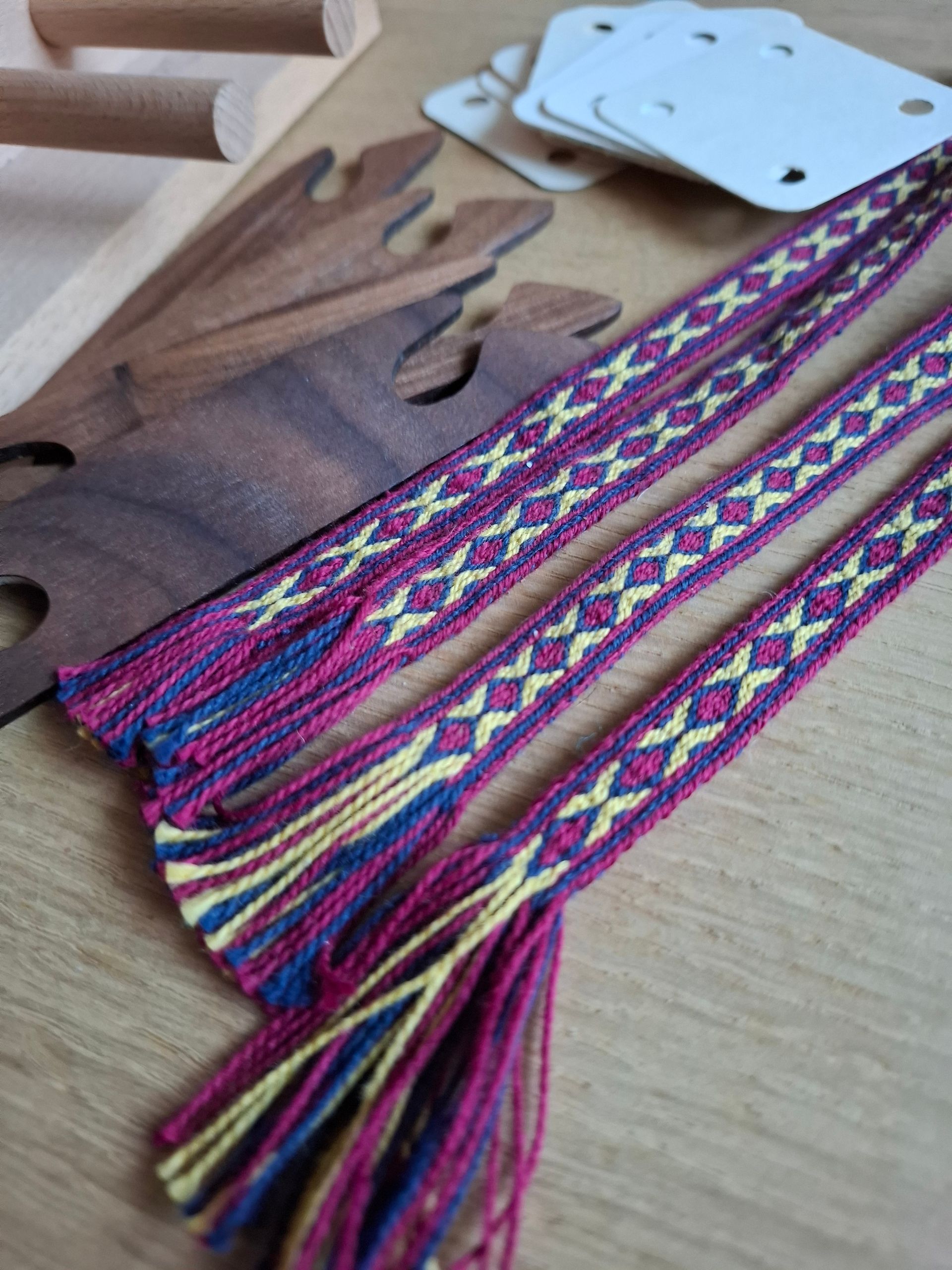 Drie geweven banden met een paars, blauw en geel patroon, met weefgereedschap op een houten oppervlak.