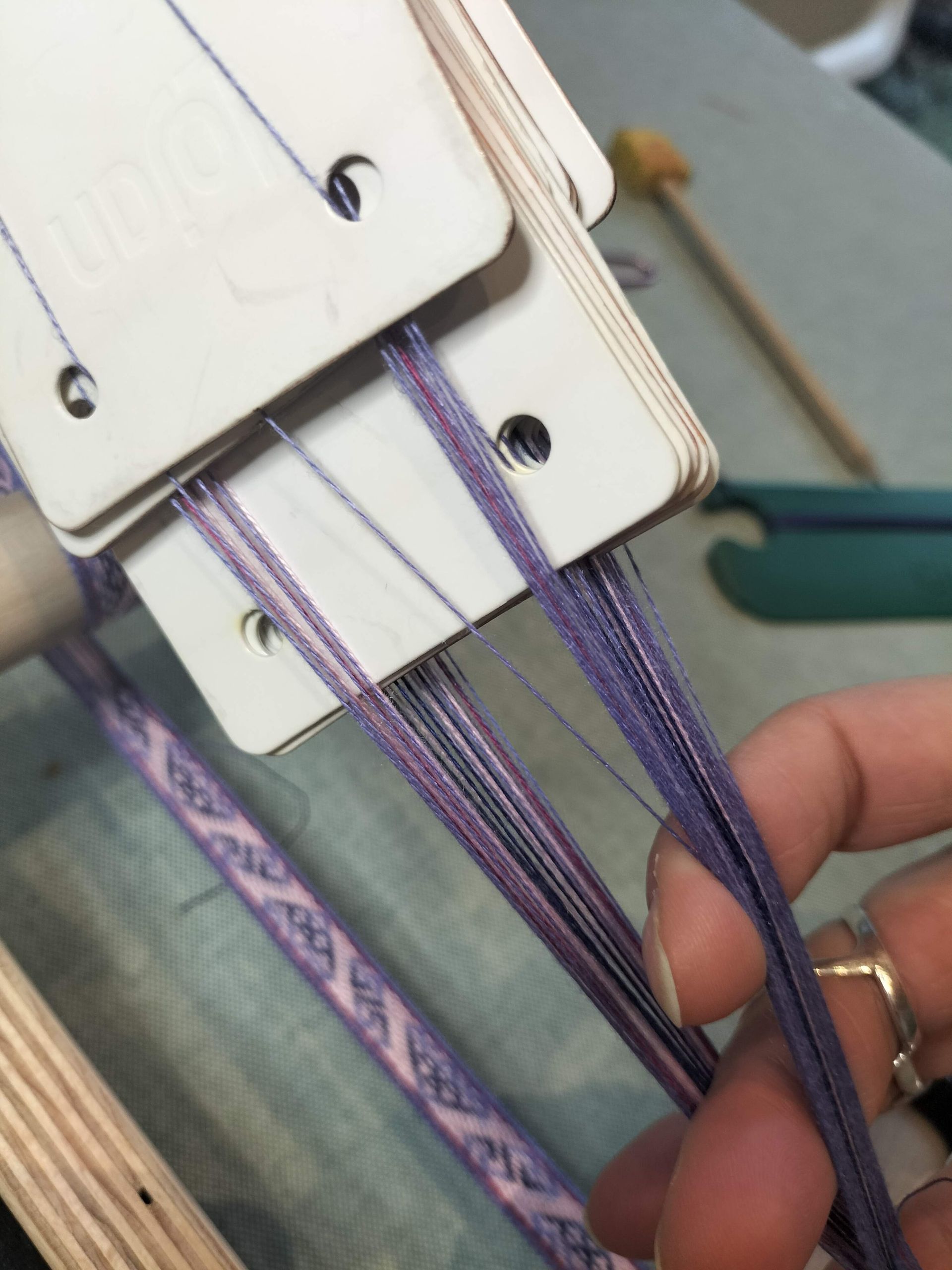 Close-up van een hand die paarse en zilveren draden door een kaartweefopstelling manipuleert.