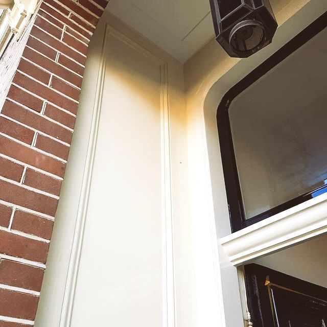 Hoek van een gebouwingang. Beige lambrisering naast rode bakstenen. Zwart omlijst raam en lamp.