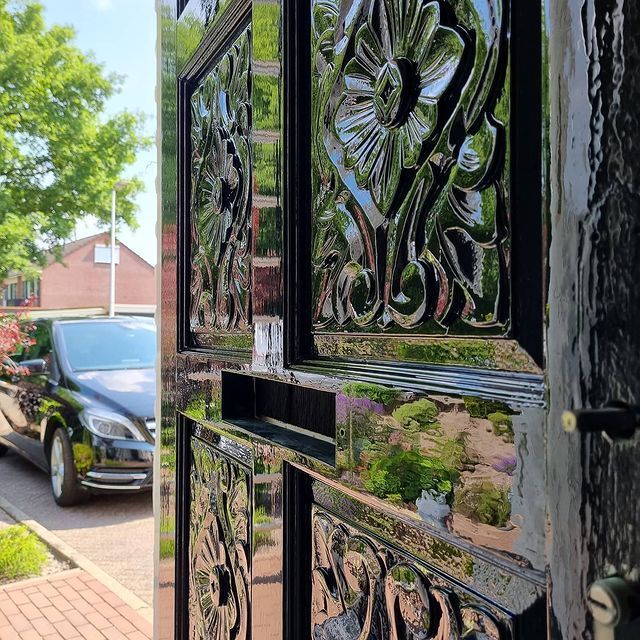 Een sierlijke zwarte deur die een auto, een huis en bomen weerspiegelt op een zonnige dag.