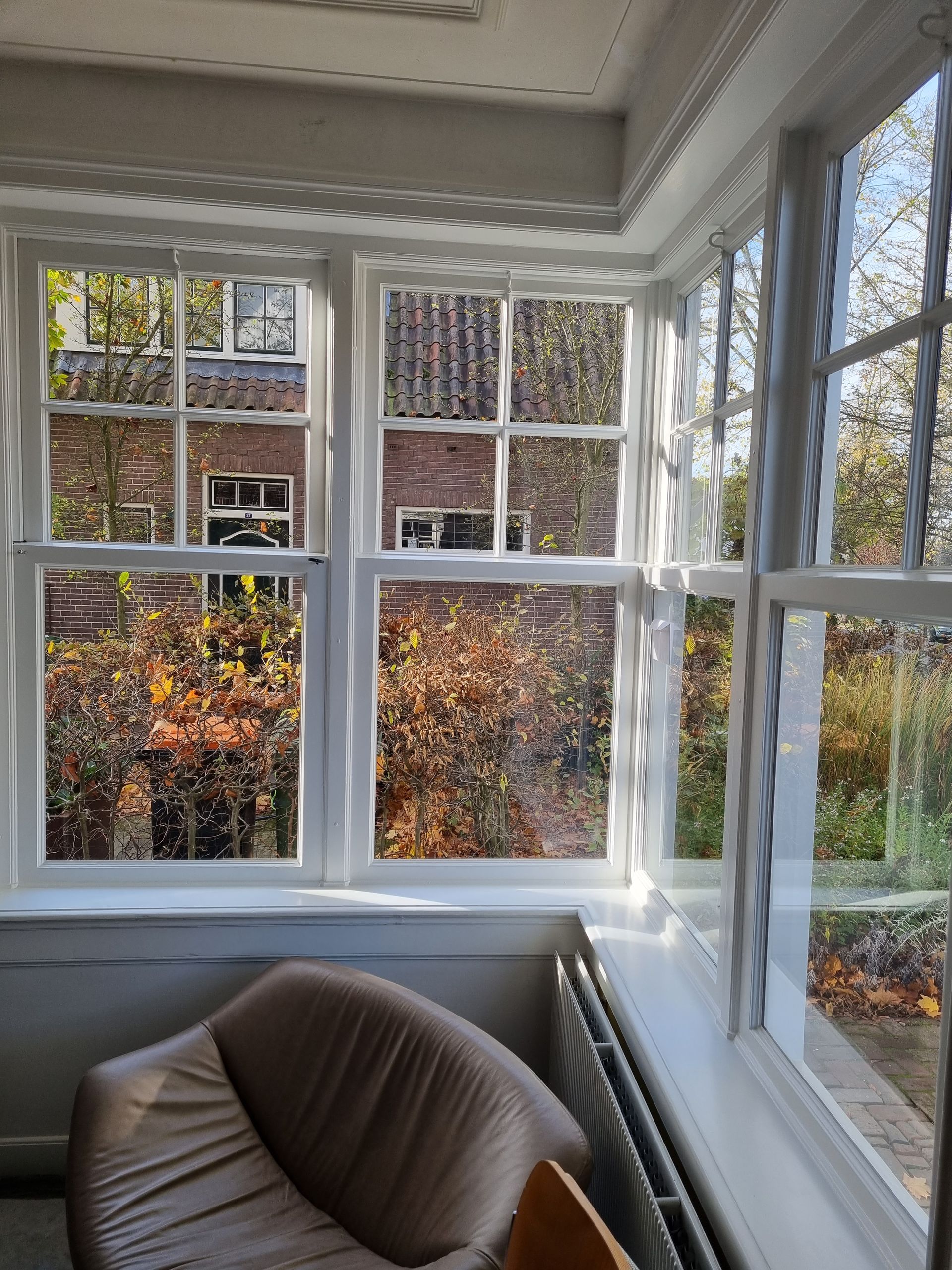 Fel zonlicht stroomt door de wit omlijste ramen en verlicht een bruine fauteuil en bomen met herfstbladeren.