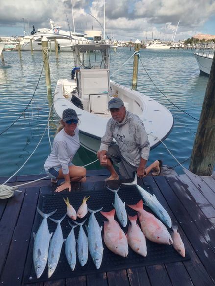 Two people pose with caught fish on a dock, boat in the background, sunny day.