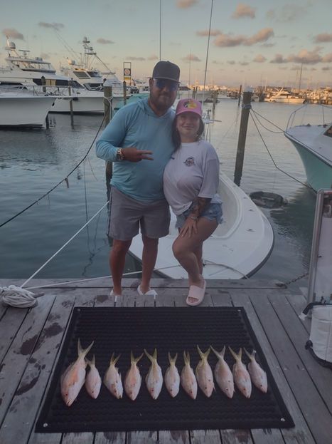 Couple posing on a dock with a row of caught fish. Boats in the background, sunset colors.
