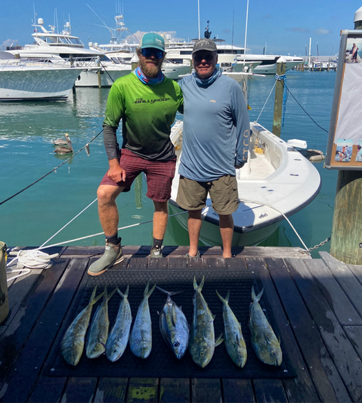 Two men pose with six fish on a dock near a boat. Yachts are in the background. Sunny day.