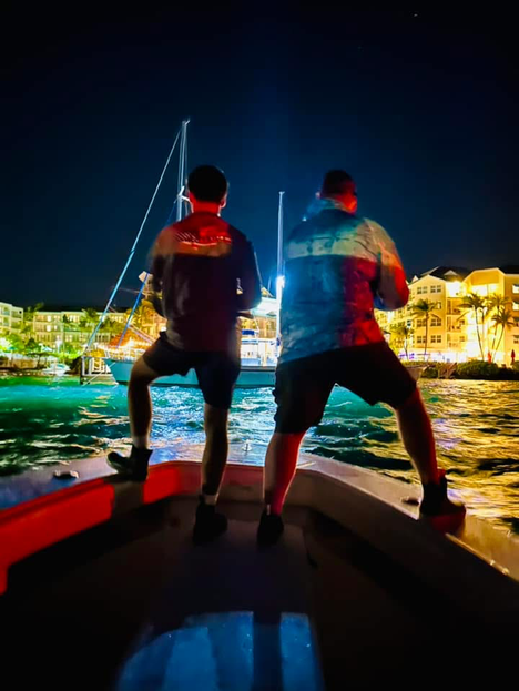 Two people on a boat at night, looking at a lit up marina.