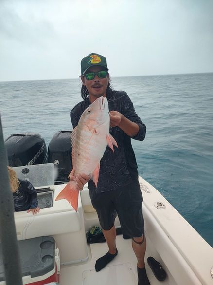 Man on boat holds up a red snapper fish. Cloudy sky, open water.