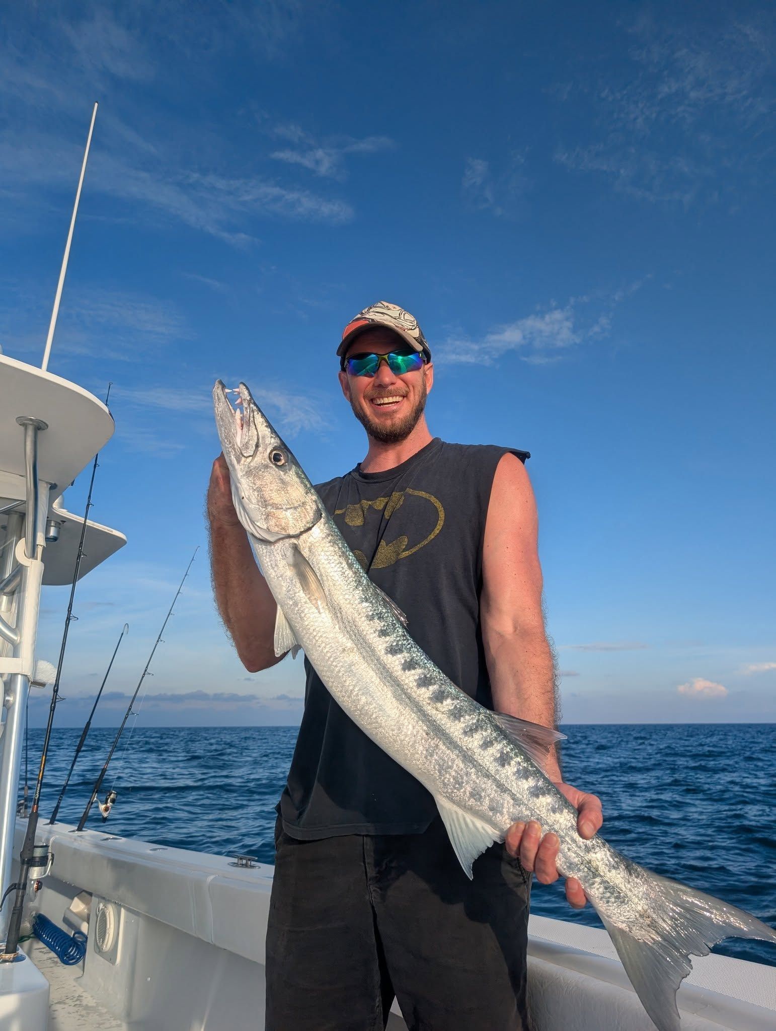 Man on boat holds a large silver barracuda fish. Blue sky and ocean in the background.
