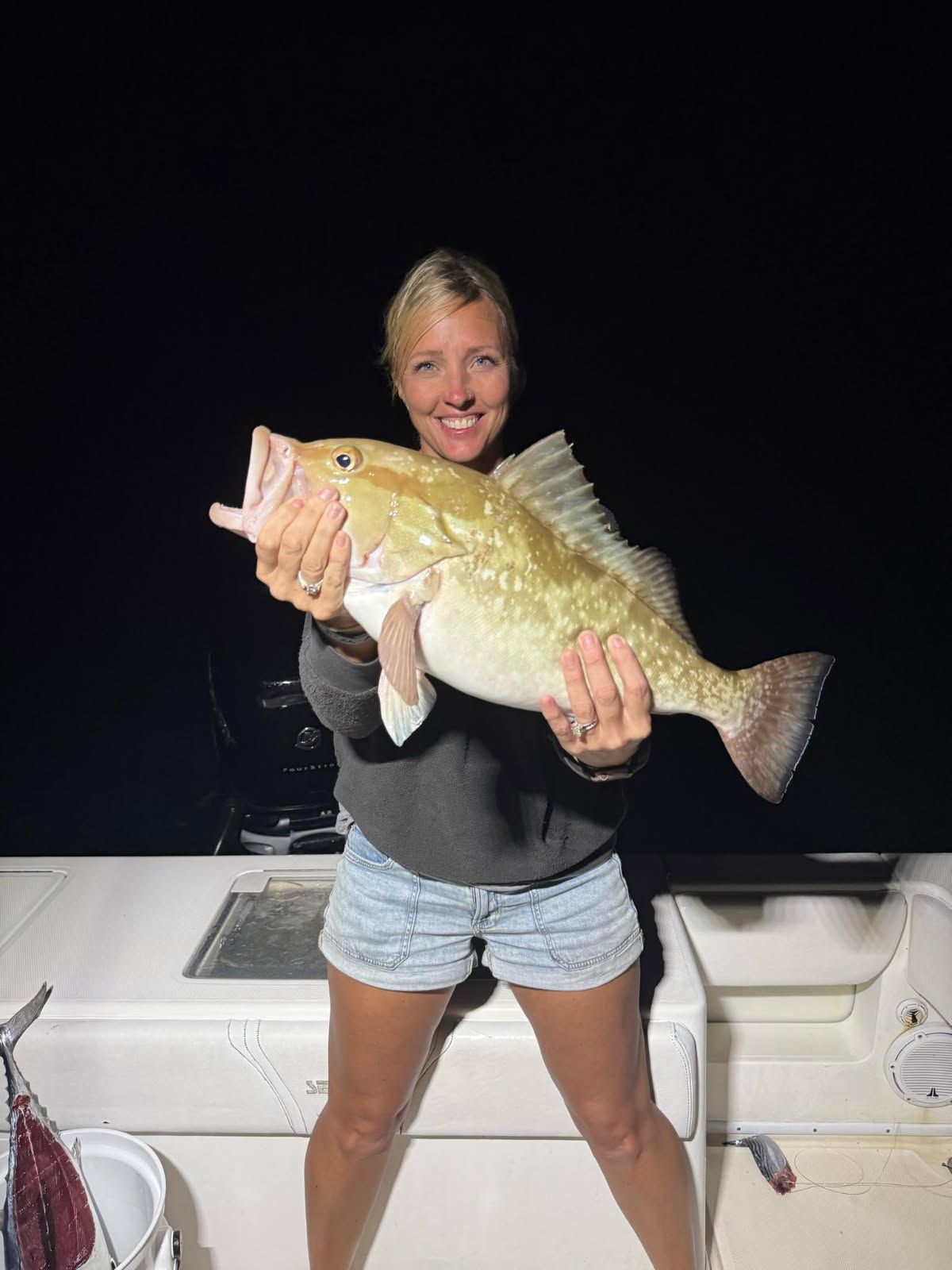Woman on a boat holding a large fish, smiling. Dark background.
