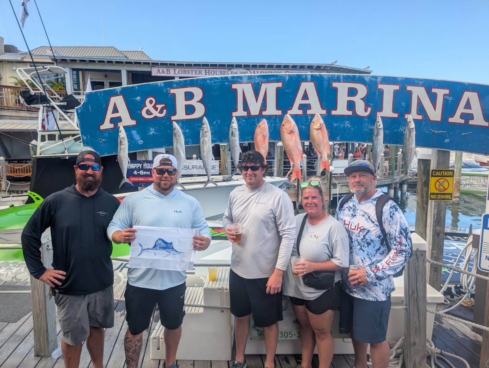 Group of people posing by A&B Marina sign with caught fish. Sunny day.