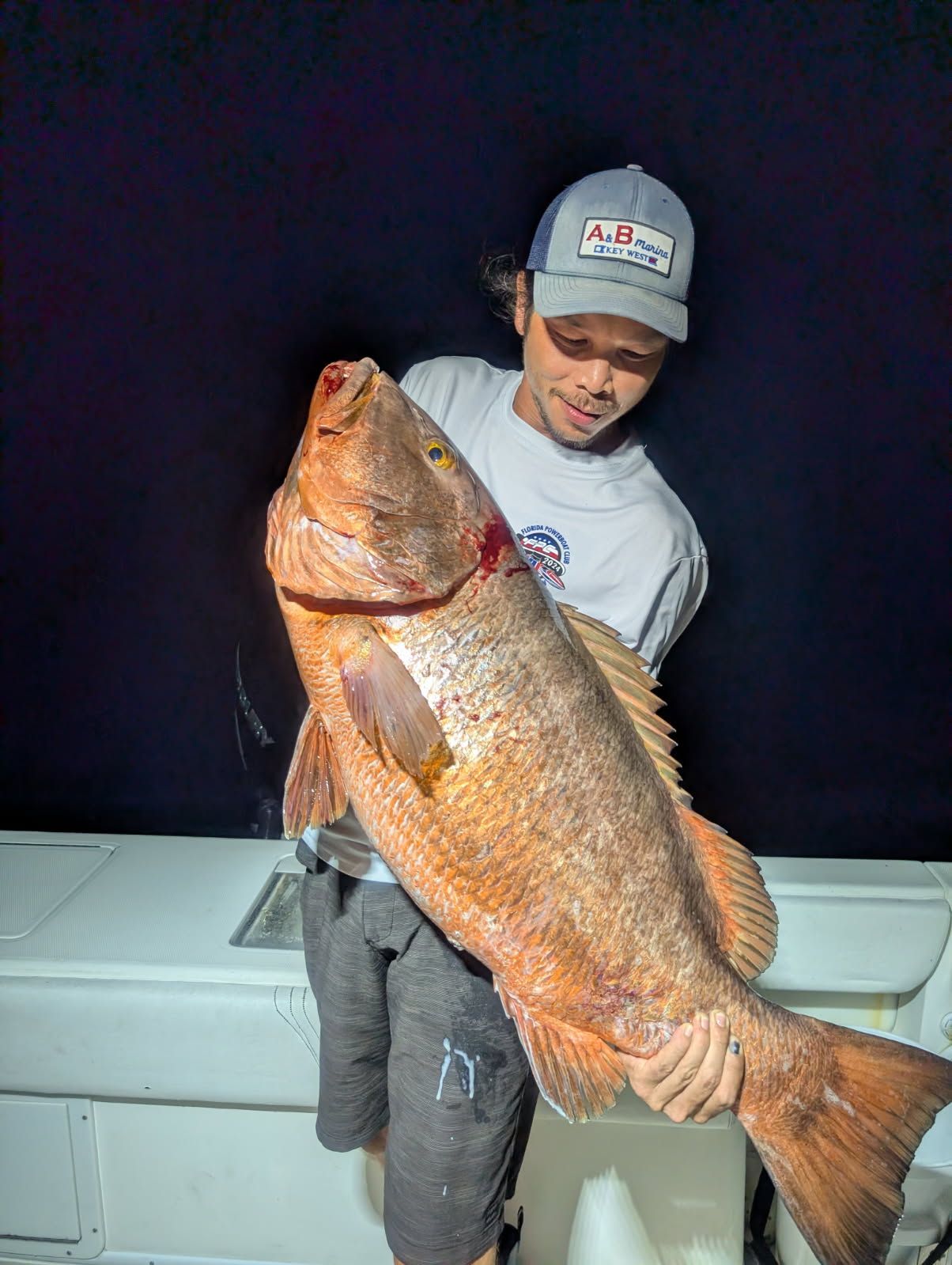 Man holding a large, reddish-brown fish on a boat at night.