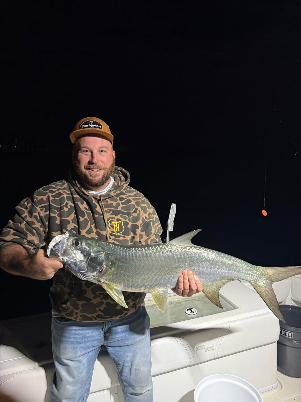 Man holding a large silver tarpon fish on a boat at night, wearing camo hoodie and hat.
