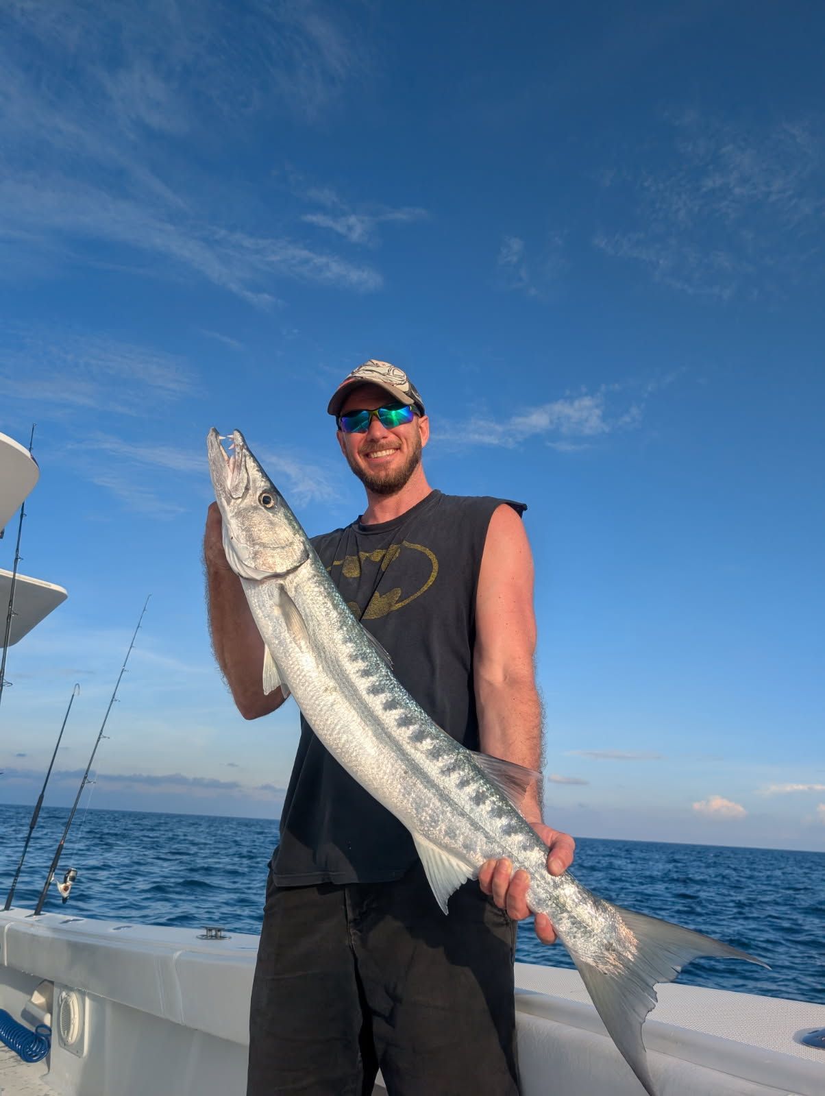 Man holding a large barracuda on a boat, blue sky, fishing rods visible.