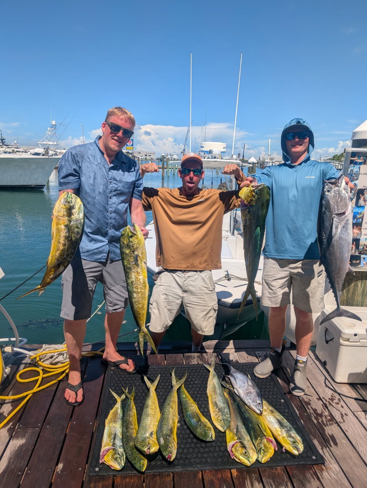Three people pose with a fishing haul: dolphin fish and a large tuna on a pier. Blue sky, harbor in background.