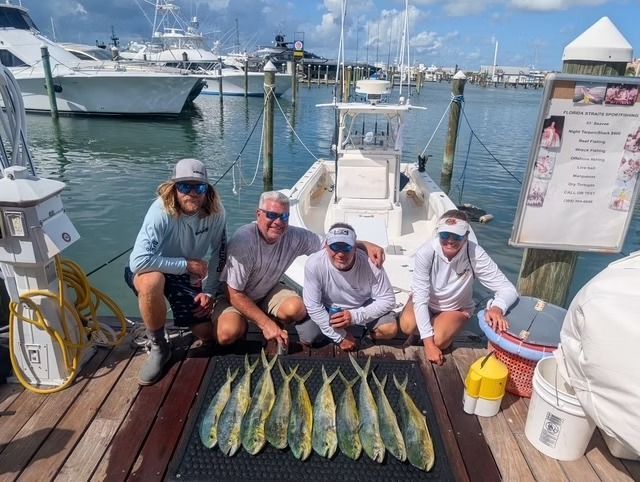Four people pose with several fish on a dock, a boat behind them in a marina setting.