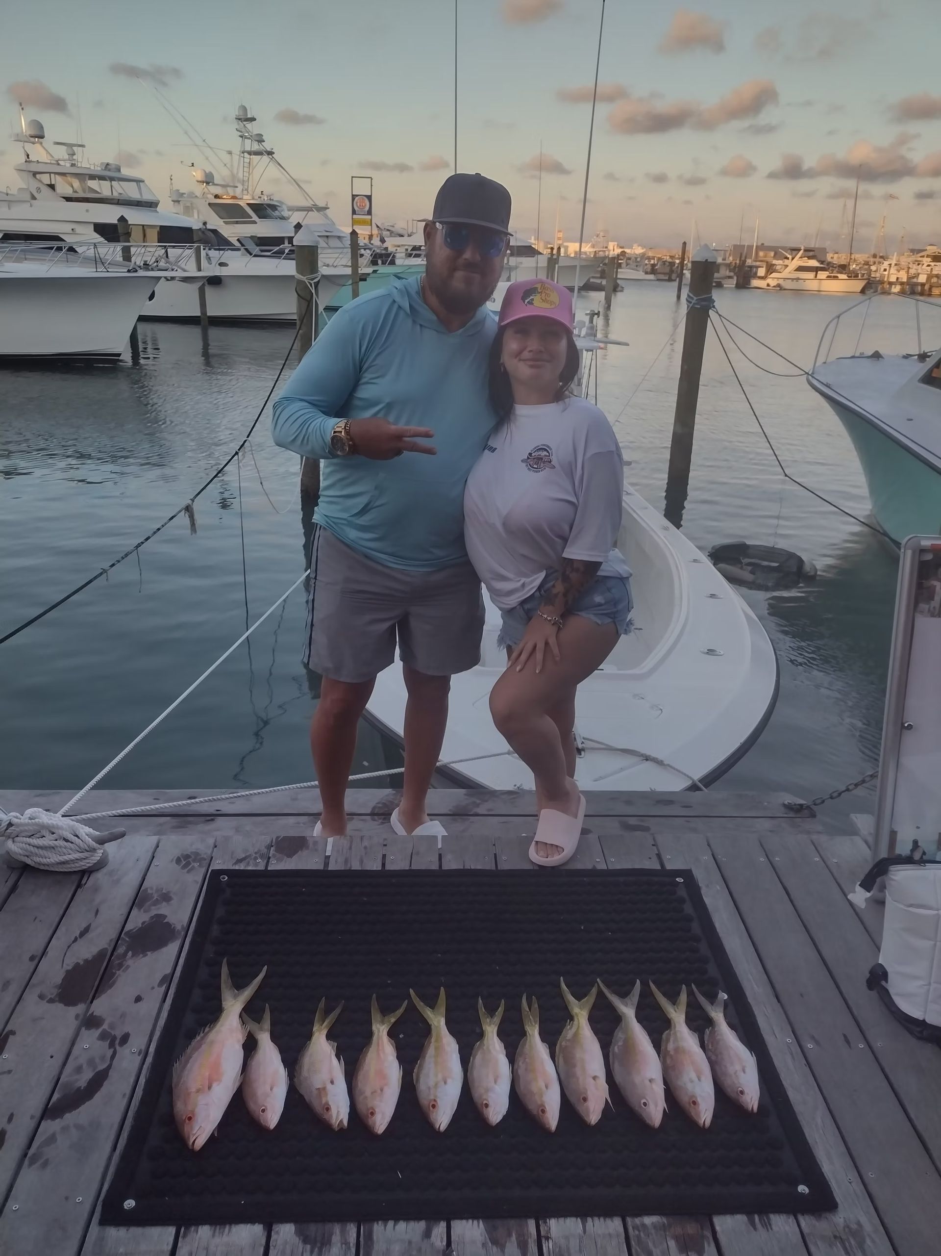 Couple poses with a row of caught fish on a pier. Boats in background.