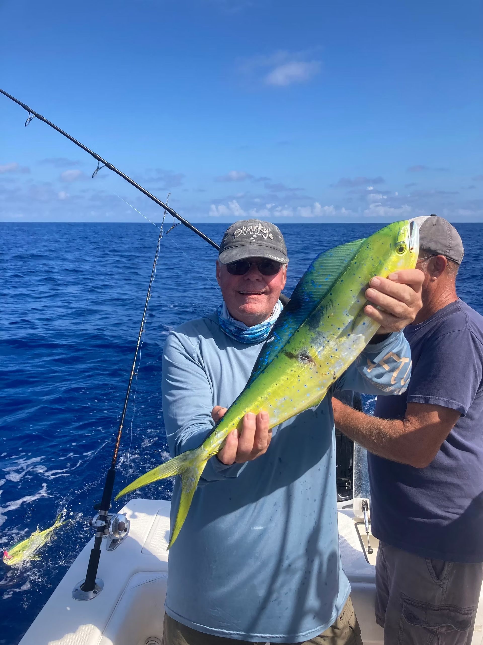 Man on boat holds up a colorful mahi-mahi fish. Another person stands beside him with ocean background.