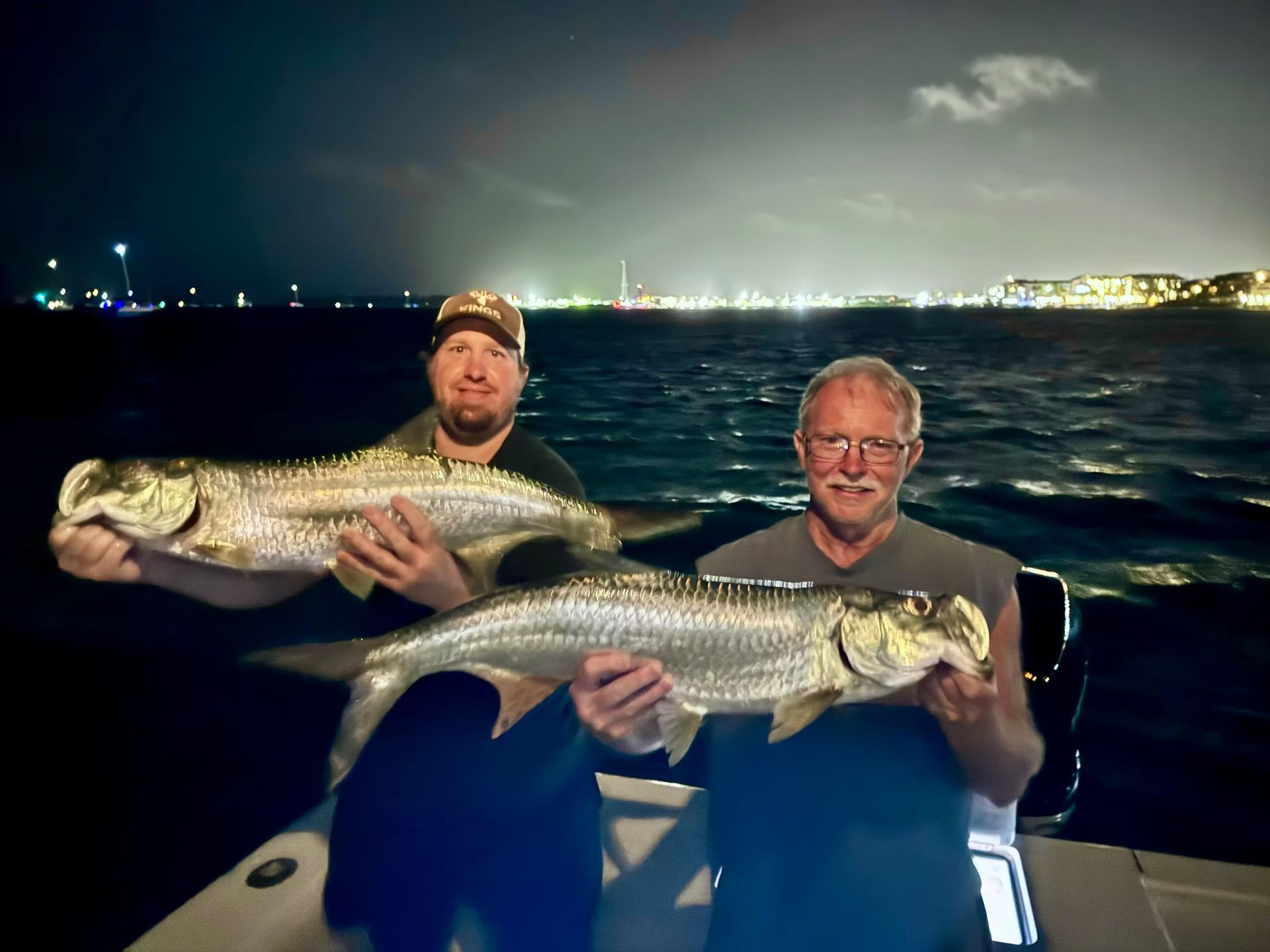 Two men on a boat at night holding large silver fish, possibly tarpon, with city lights in the background.