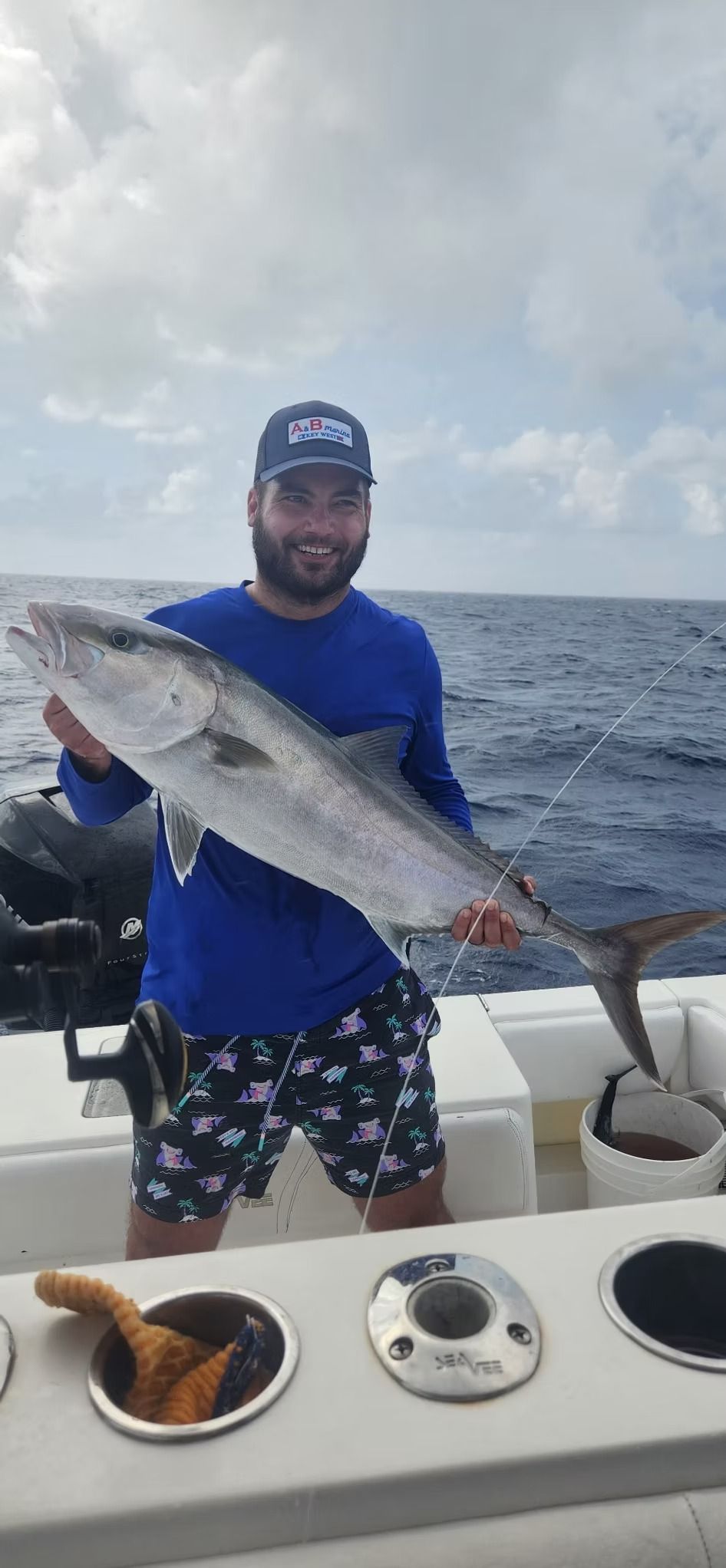 Man on a boat holding a large fish; blue shirt, patterned shorts, and hat; ocean in the background.