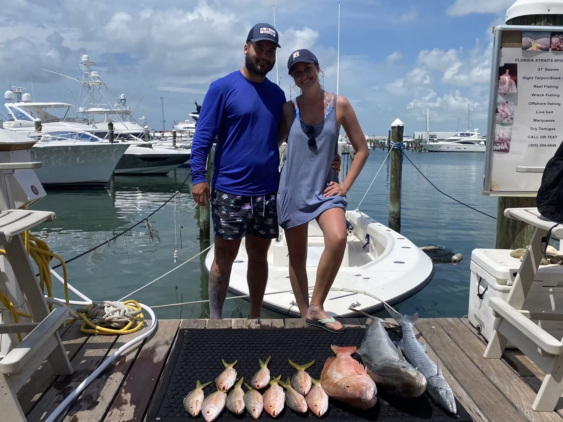 Couple poses with their catch of fish at a marina. Blue sky, water, boats.
