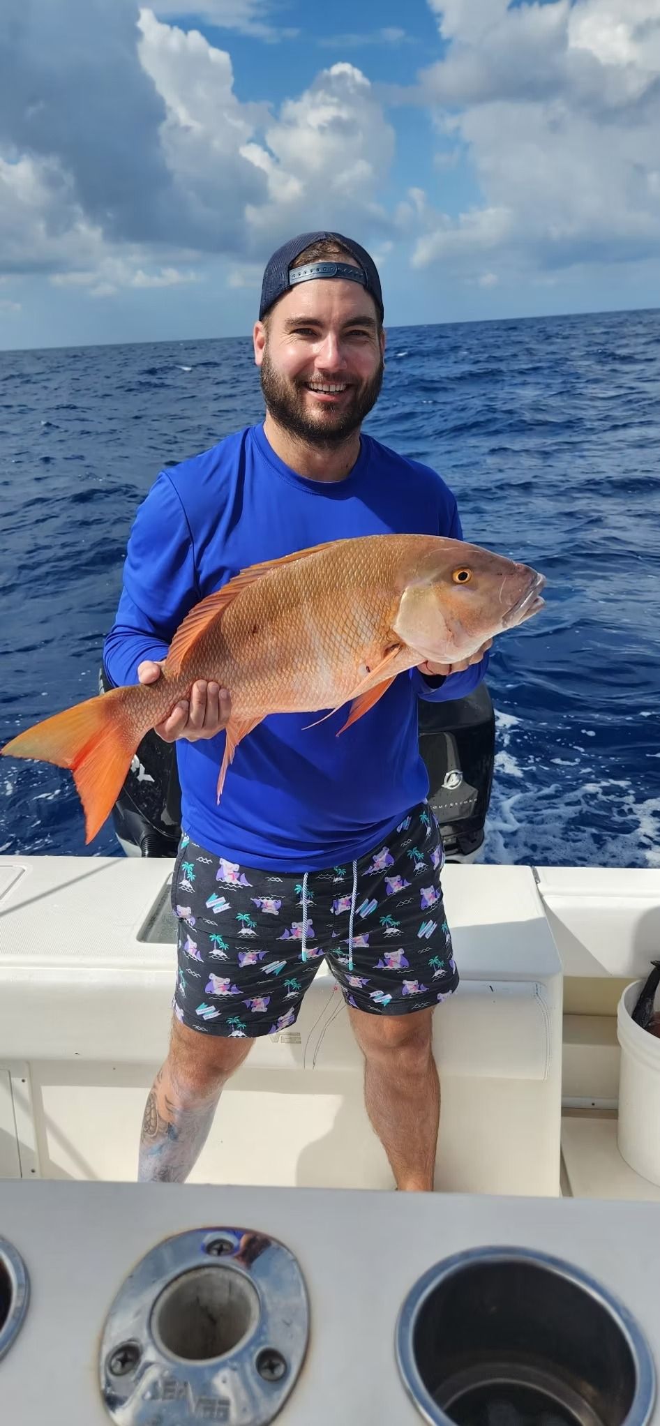 Man holding large fish on a boat; blue shirt, patterned shorts, smiling; ocean background.