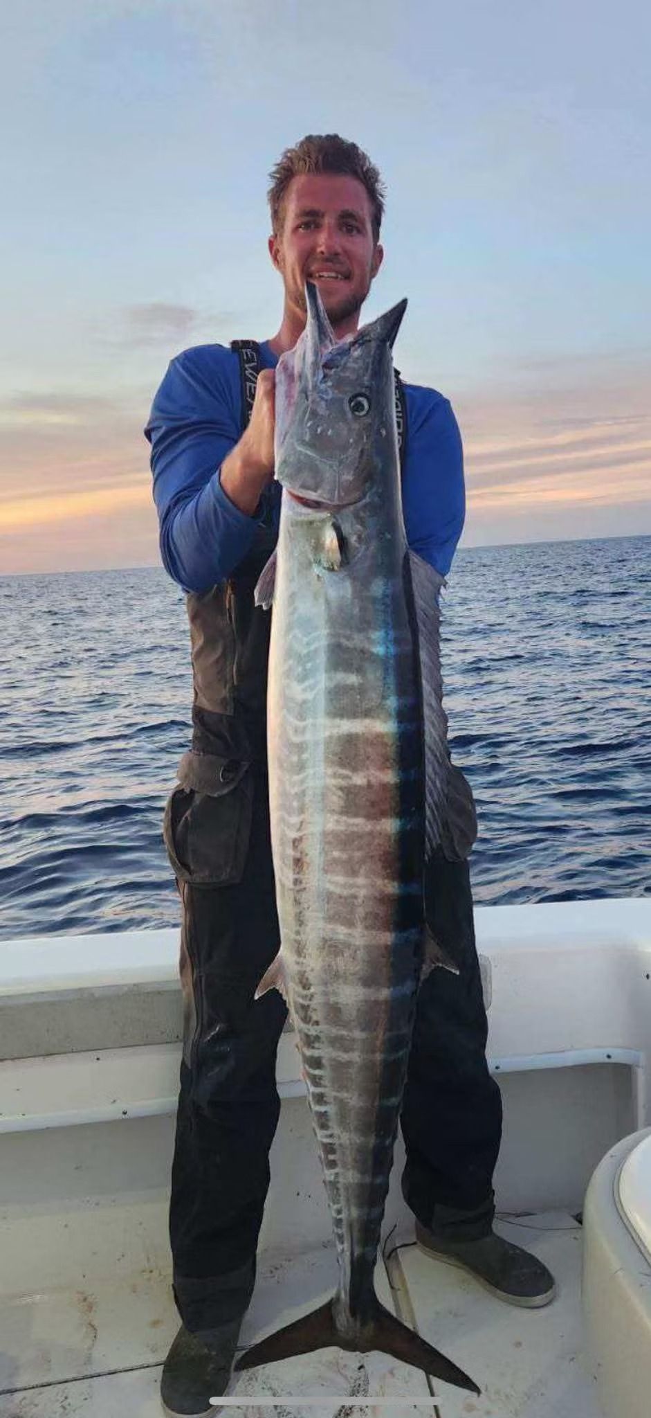 A person holding a large fish on a boat. The man wears blue shirt and dark pants. Sky and water in background.