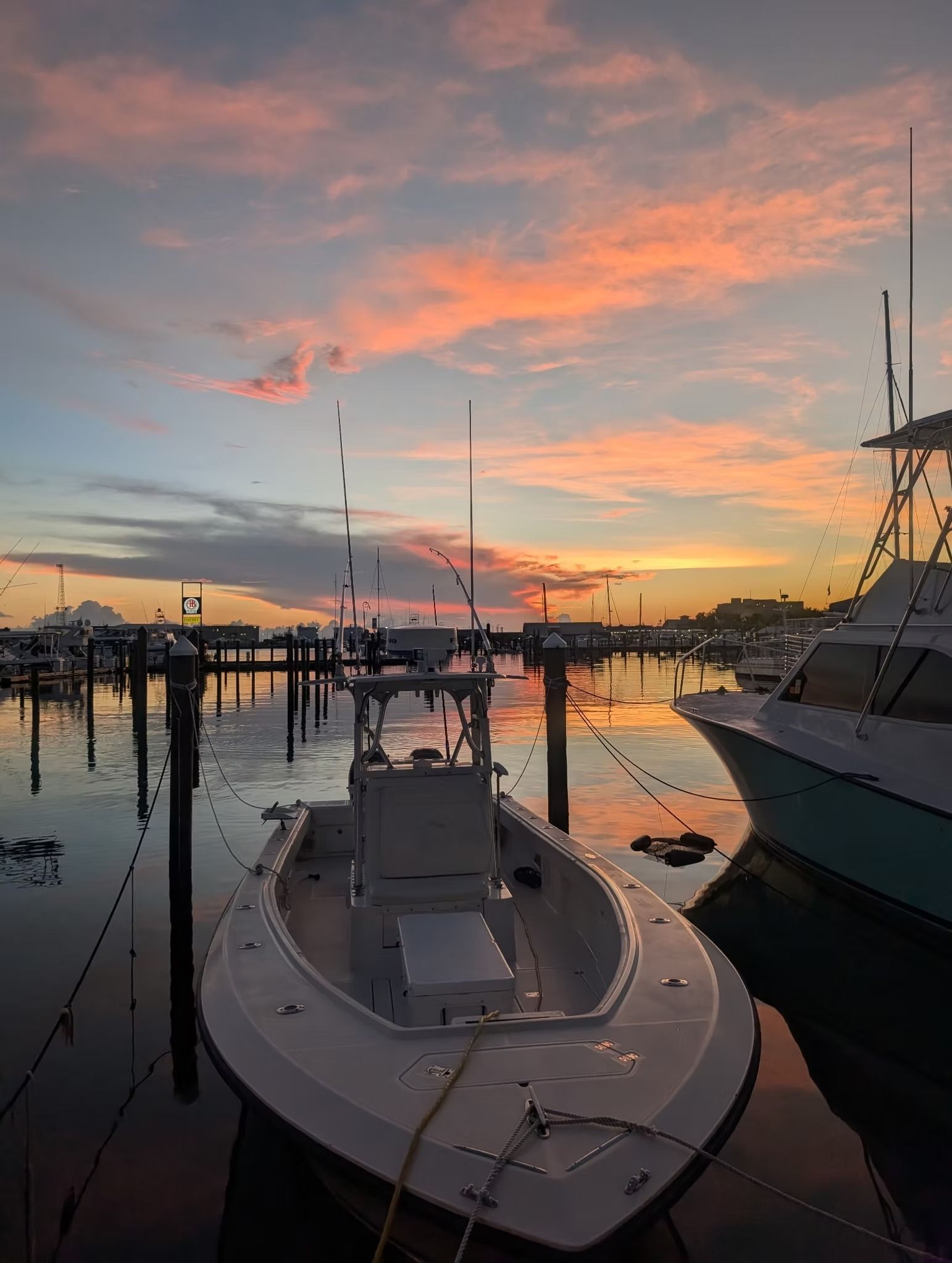 White boat docked at harbor at sunset; orange and pink sky reflects on calm water.