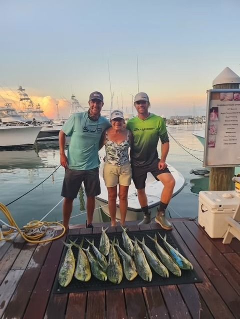 Three people posing with a row of caught fish on a dock at sunset, with boats in the background.