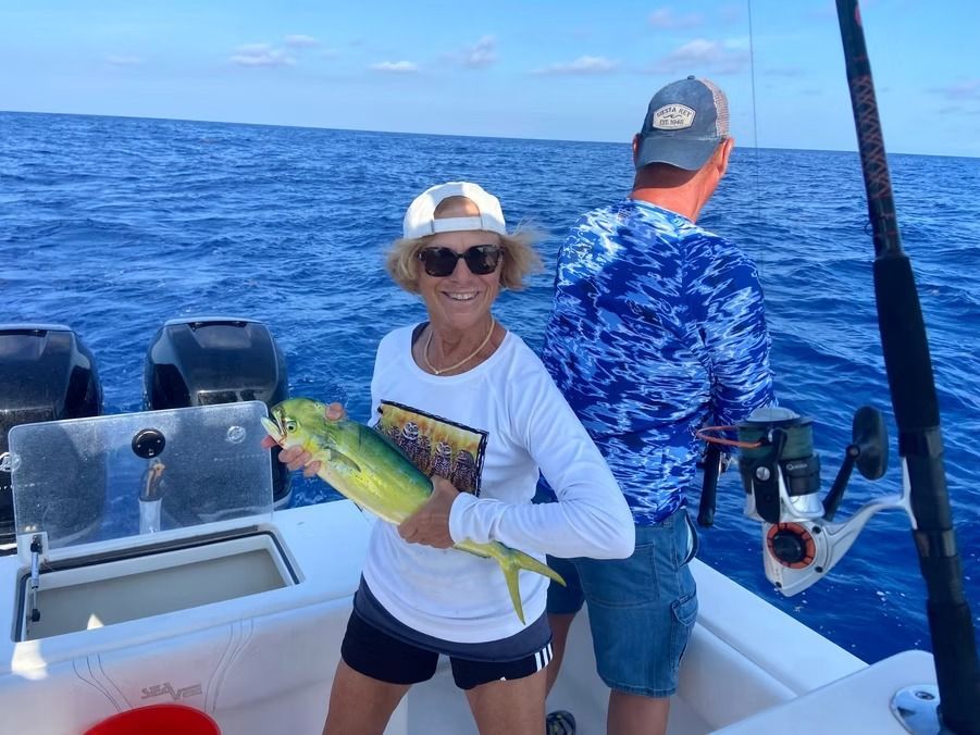 Woman on a boat holding a fish she caught, the man behind her is fishing too, on the ocean.