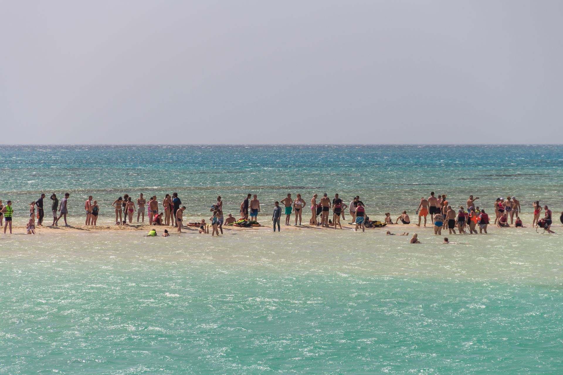 People standing and walking on a sandbar surrounded by turquoise water, under a bright sky.