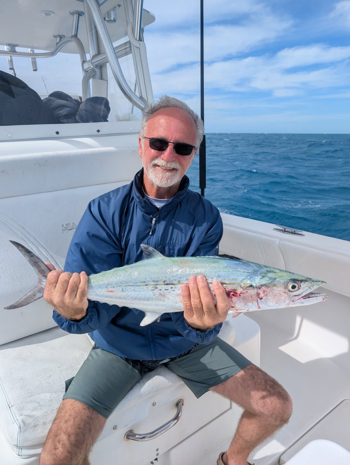 Man on a boat holding a fish he caught, smiling. Blue ocean and sky in background.