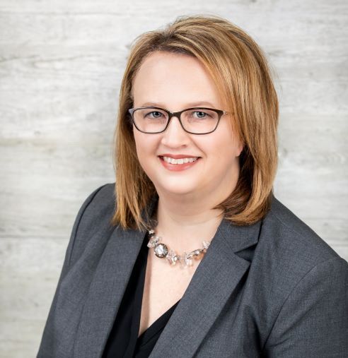 Woman with glasses wearing a gray blazer smiles at the camera, posing against a light background.