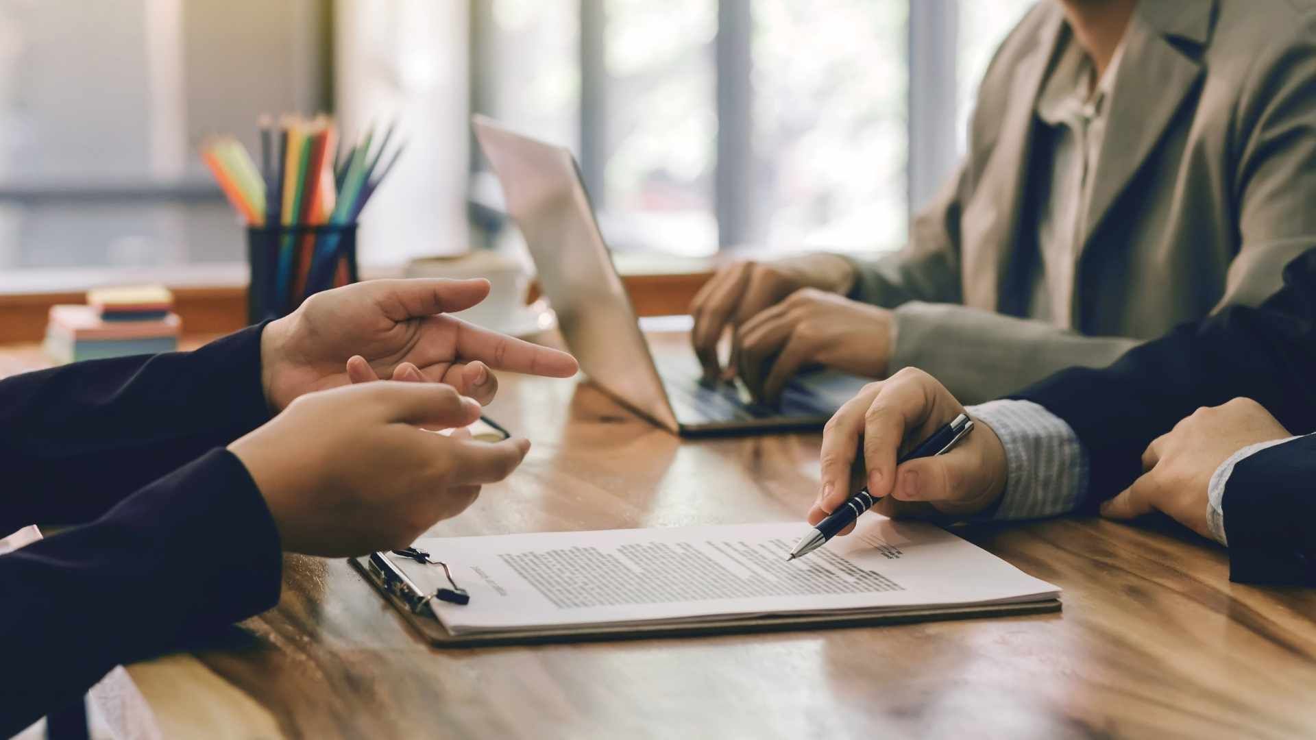 People in suits reviewing a document, with a laptop and office supplies on a wooden table.